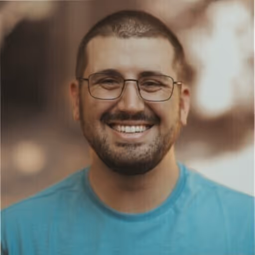 Smiling man with short hair, beard, glasses, and a blue shirt against a blurred background.