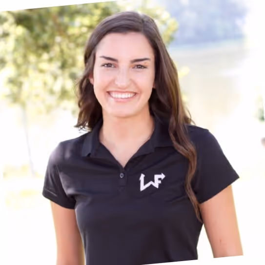 Smiling woman with long brown hair wearing a black polo shirt with a white logo, standing outdoors with blurred greenery and water in the background.