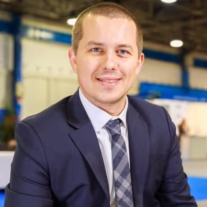 Smiling man in a dark suit and patterned tie sitting indoors with a blurred background.