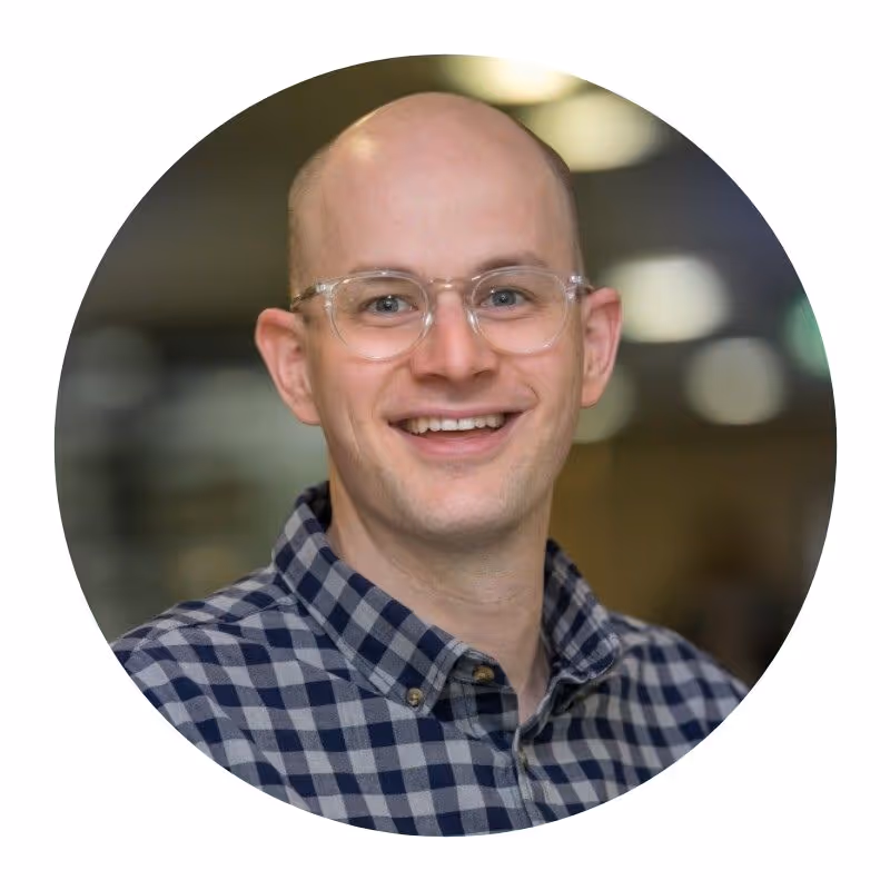 Smiling bald man wearing clear glasses and a blue checkered shirt in an indoor setting.