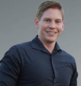 Smiling man with short light brown hair wearing a dark blue checkered shirt against a plain gray background.