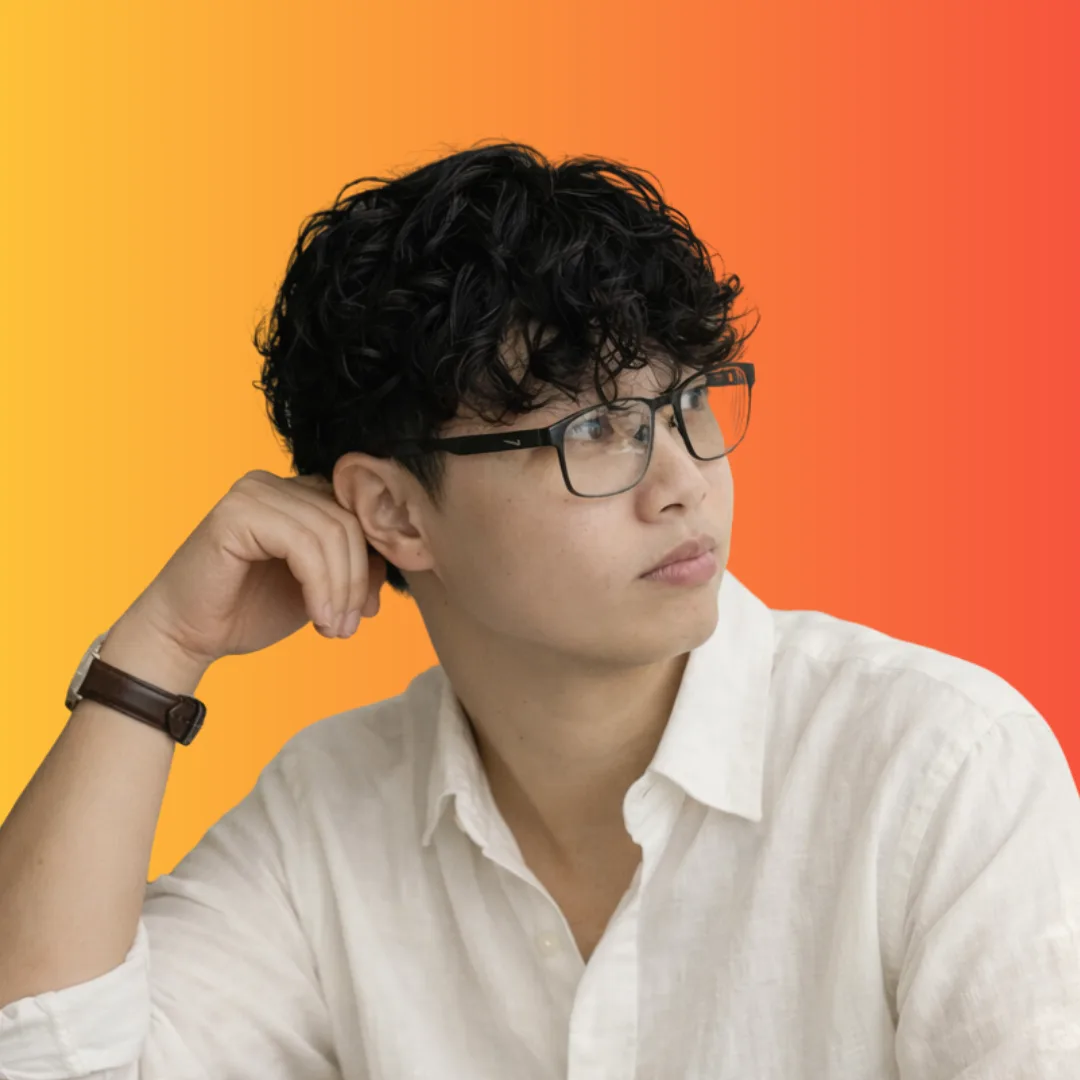 Woman with dark curly hair and black-framed glasses in a white shirt, hand raised to the side of her head, against an orange to red gradient background.