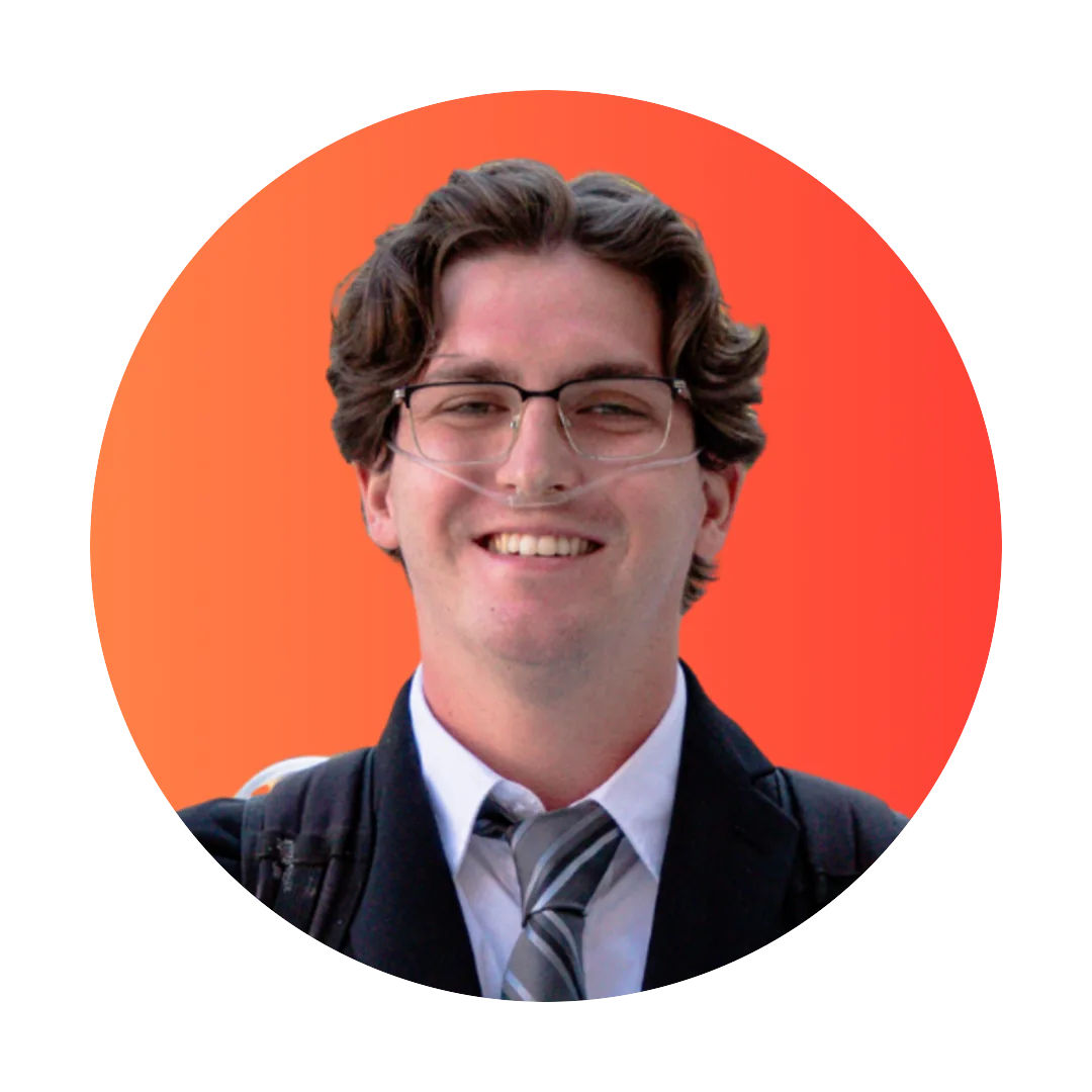 Smiling young man with wavy brown hair and clear-framed glasses wearing a dark suit jacket, white shirt, and gray striped tie on an orange to red circular background.