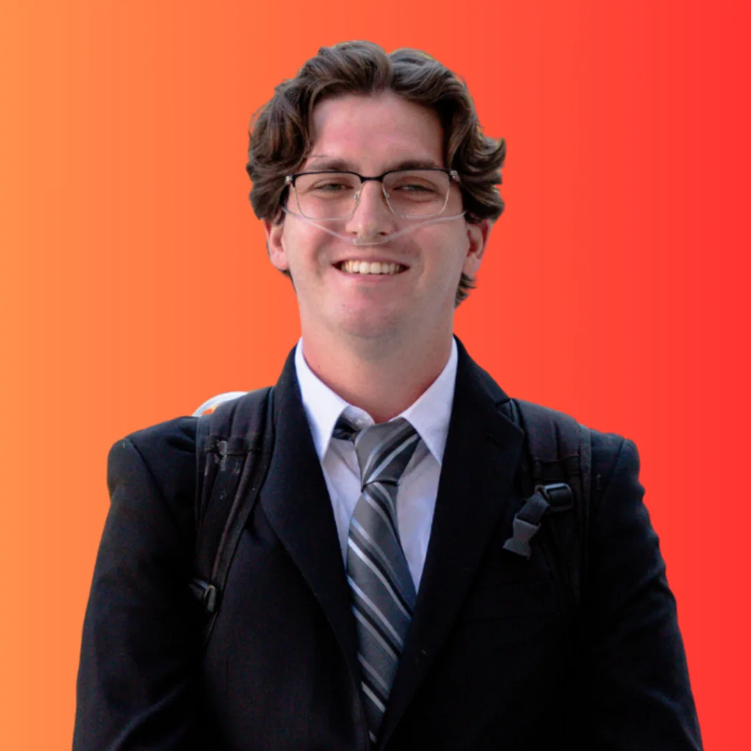 Smiling young man with wavy brown hair and clear-framed glasses wearing a dark suit jacket, white shirt, and gray striped tie with backpack straps over his shoulders, against an orange to red gradient background.